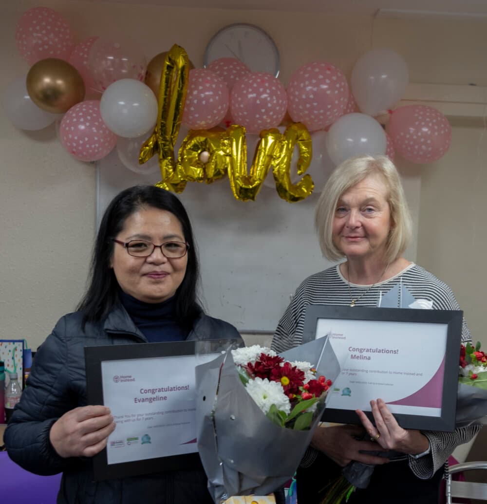 Two women holding certificates and bouquets stand in front of a "Congratulations" balloon and pink and gold balloons. - Home Instead