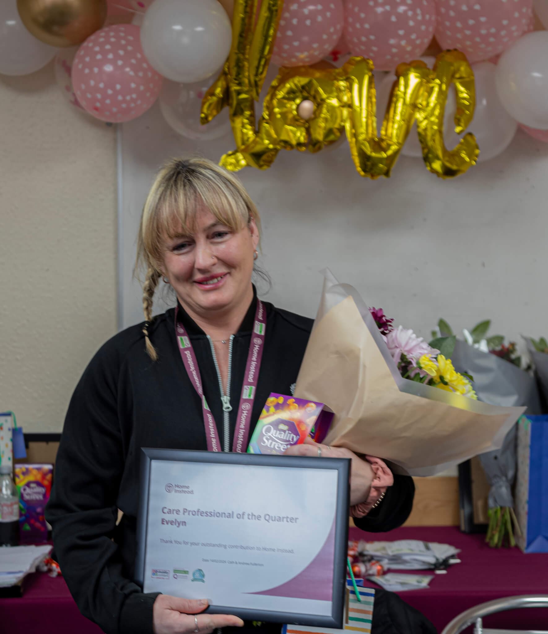 A woman holds an award and bouquet, standing in front of balloons spelling "love. - Home Instead