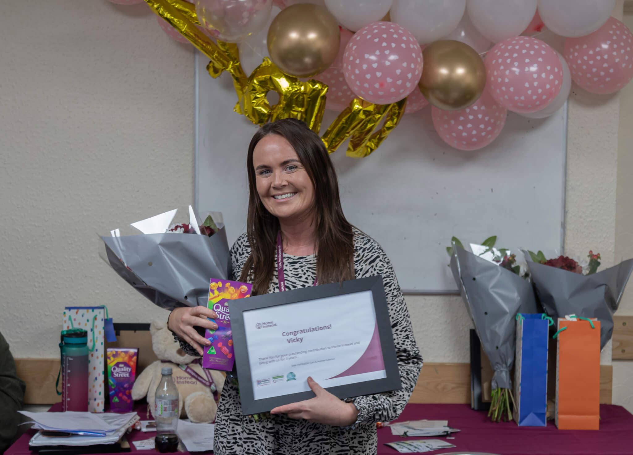 A woman smiles holding a certificate and chocolates, standing in front of balloons and gifts. - Home Instead