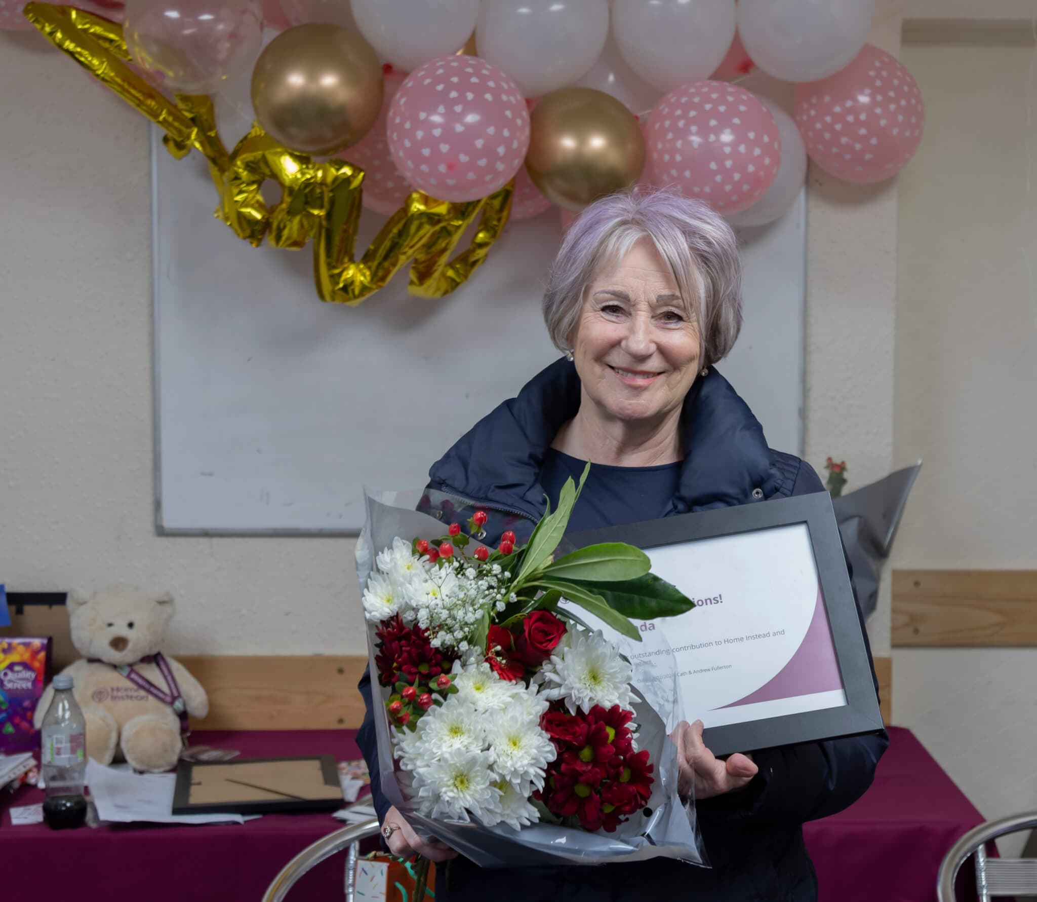 A woman holding a bouquet of flowers and a framed certificate, smiling, with balloons and gifts in the background. - Home Instead