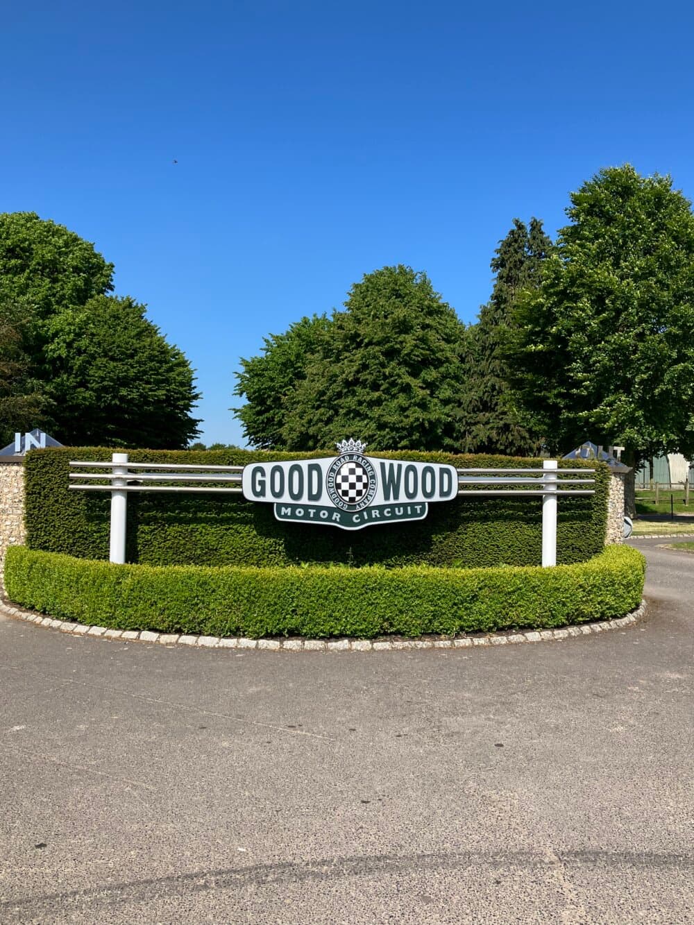 Sign for Goodwood Motor Circuit surrounded by manicured hedges and trees against a clear blue sky. - Home Instead
