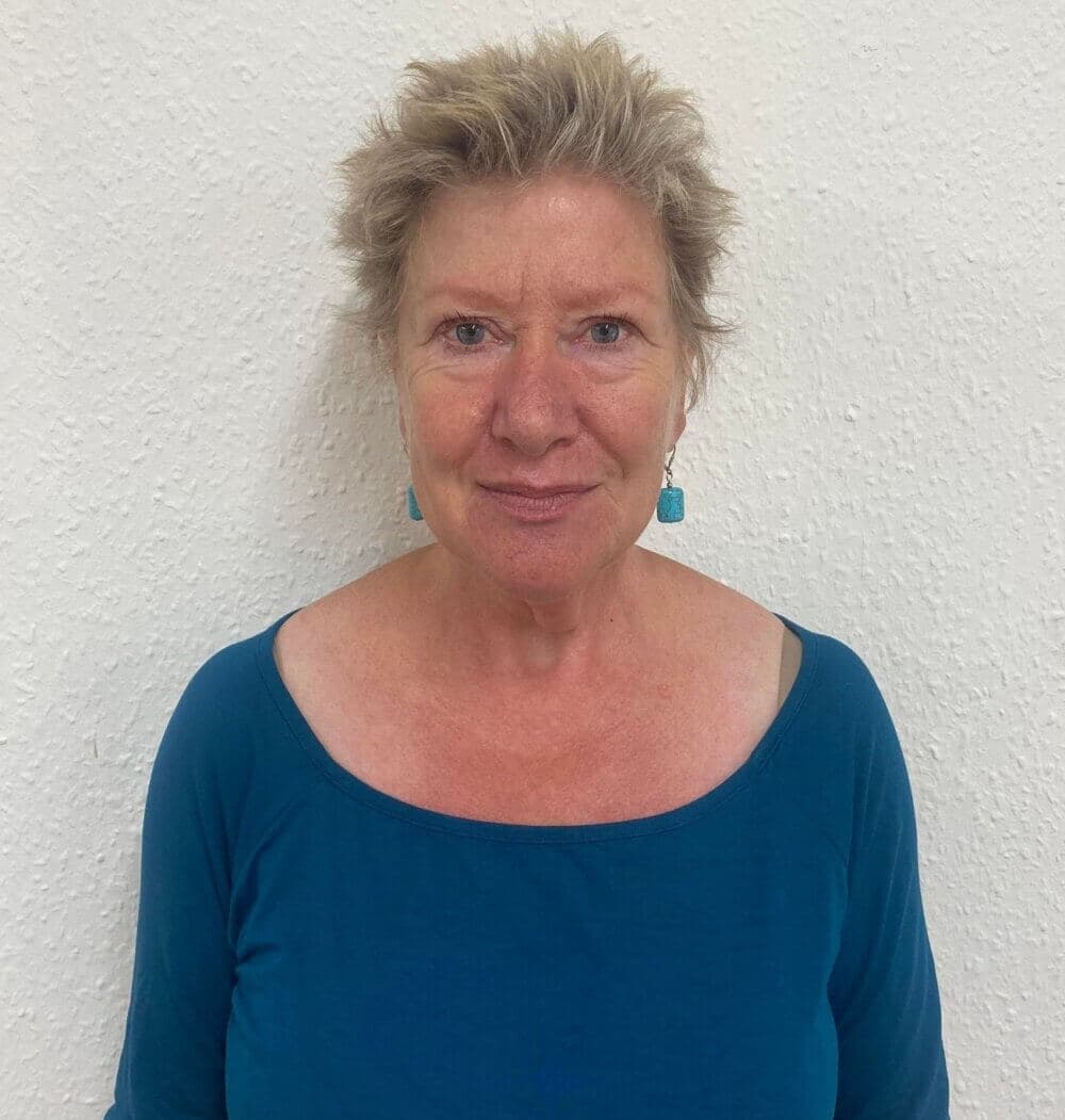 A woman with short, blond hair wearing a blue top and turquoise earrings stands against a textured white wall. - Home Instead