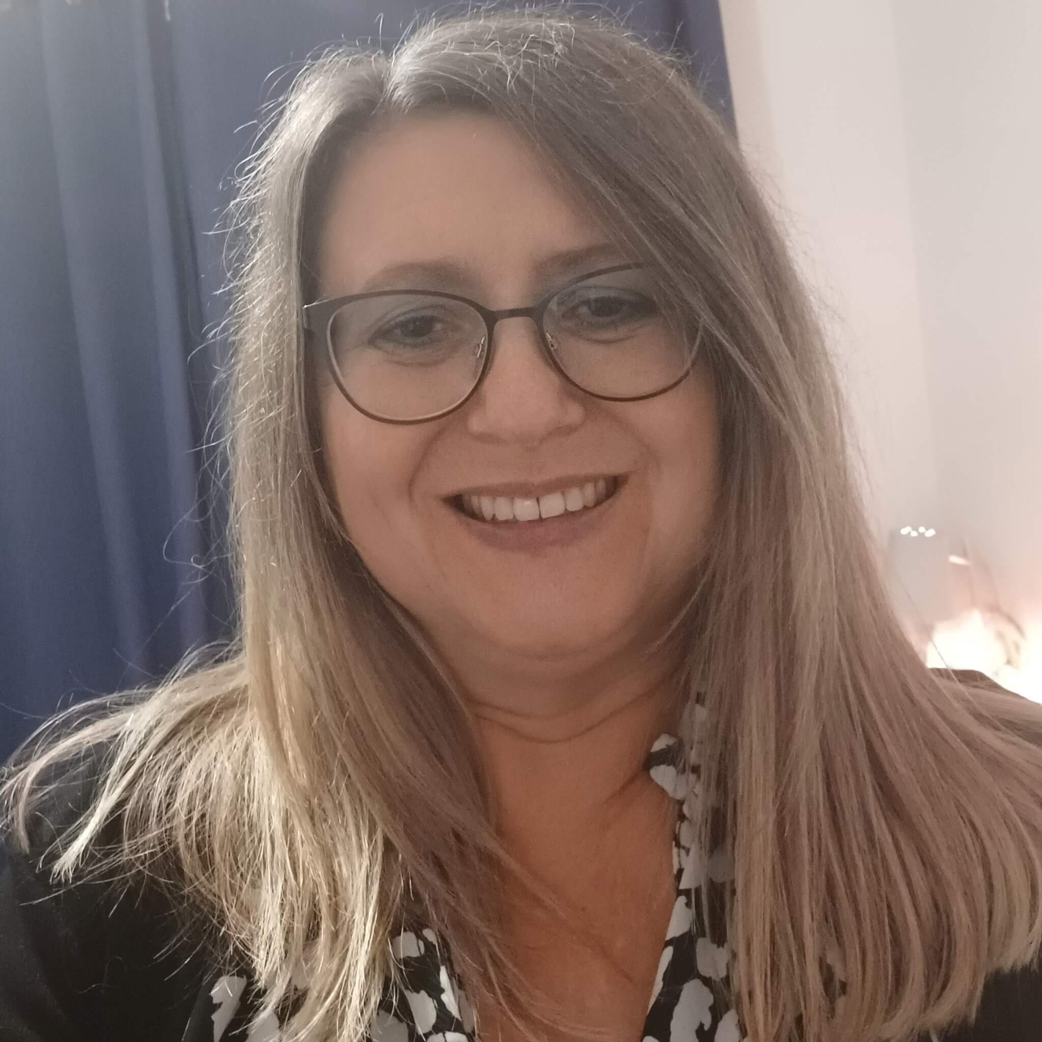 A woman with glasses and long, straight hair smiles at the camera, with dark curtains and a lamp in the background. - Home Instead