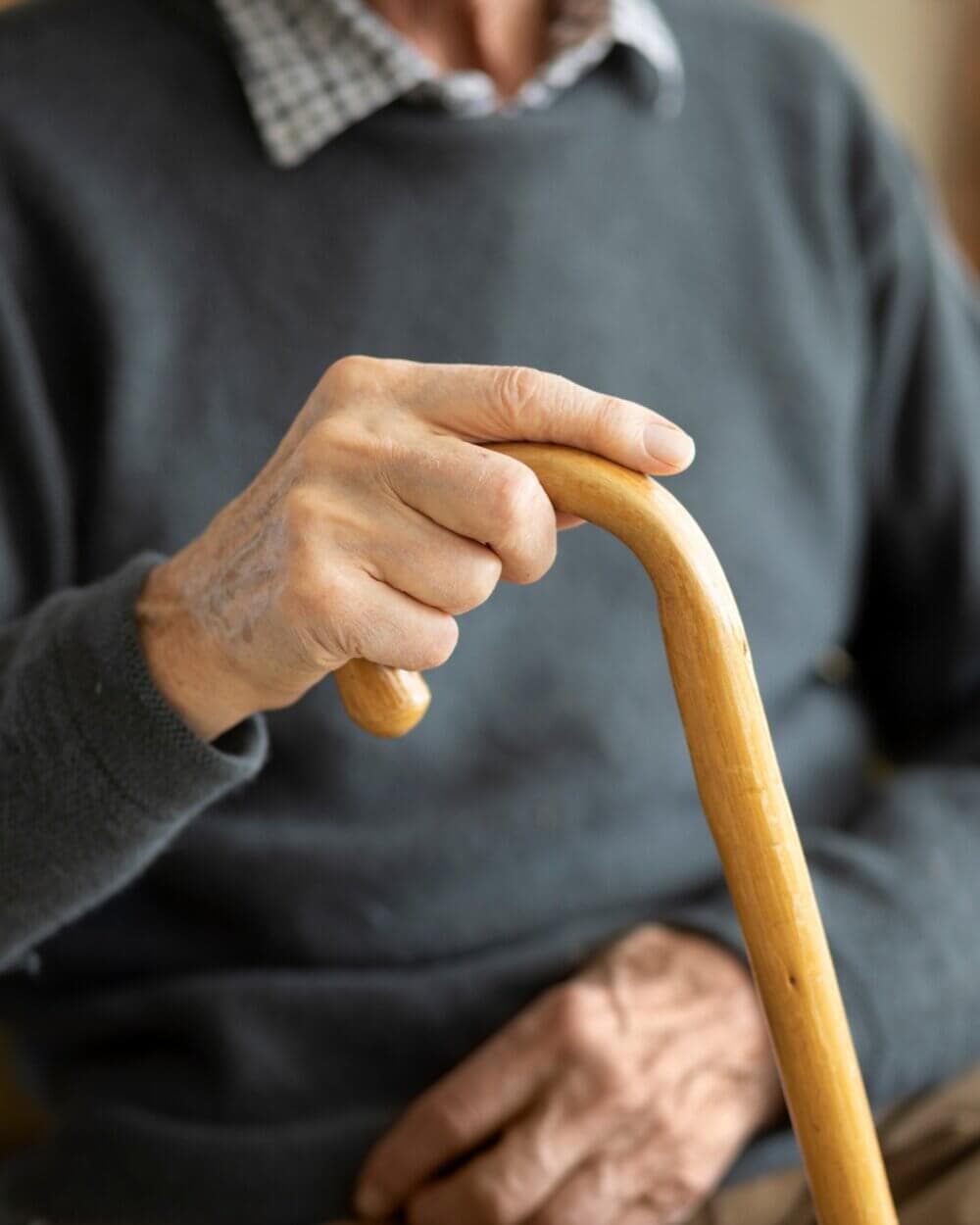 An elderly person in a dark sweater holds a wooden cane and rests their other hand on their lap. - Home Instead