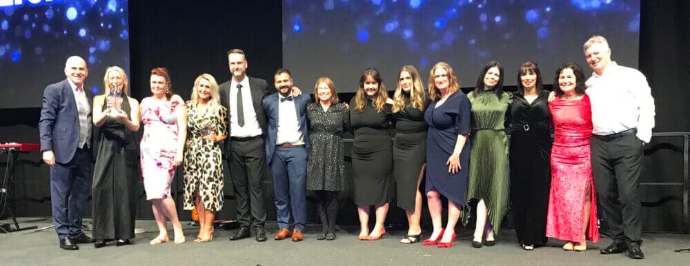 A group of people standing side by side on stage, dressed in formal attire, smiling at the camera. - Home Instead