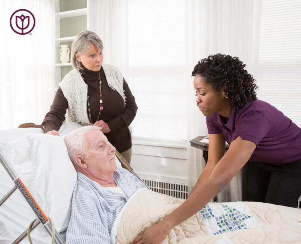 A caregiver assists an elderly man in bed while a concerned woman stands by his side in a brightly lit room. - Home Instead