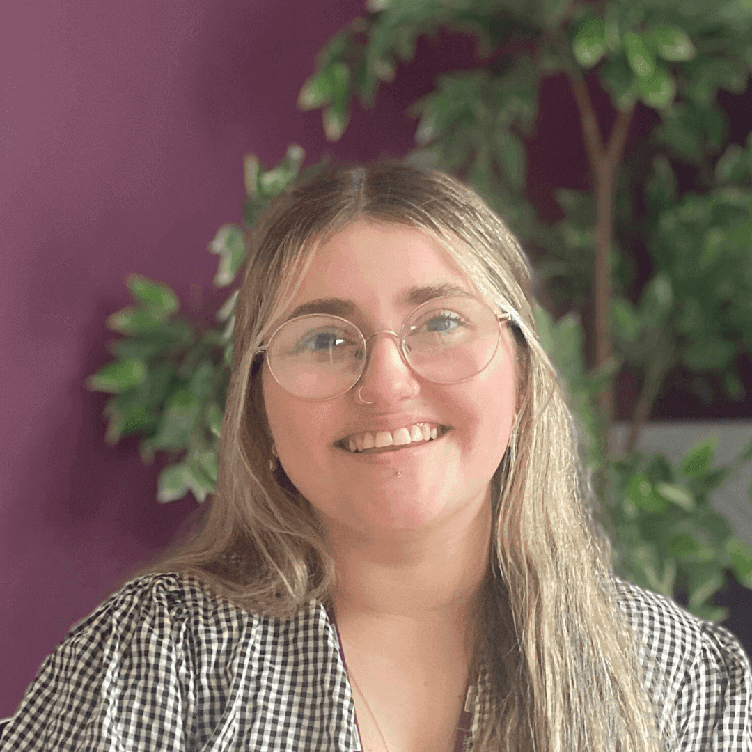 A woman with glasses smiles in front of a purple wall and a leafy plant. - Home Instead
