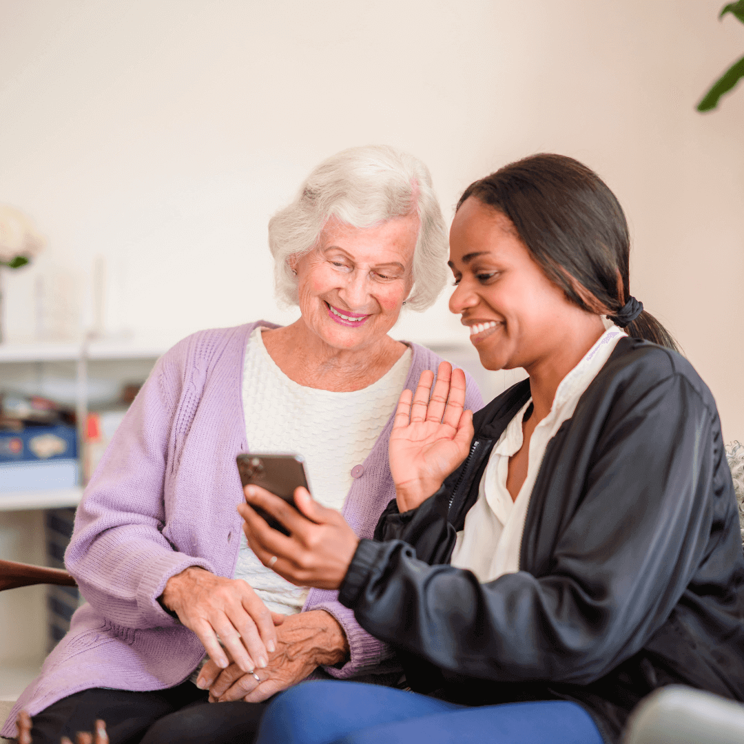 An elderly woman and a young woman smile while looking at a smartphone; the young woman waves at the phone. - Home Instead