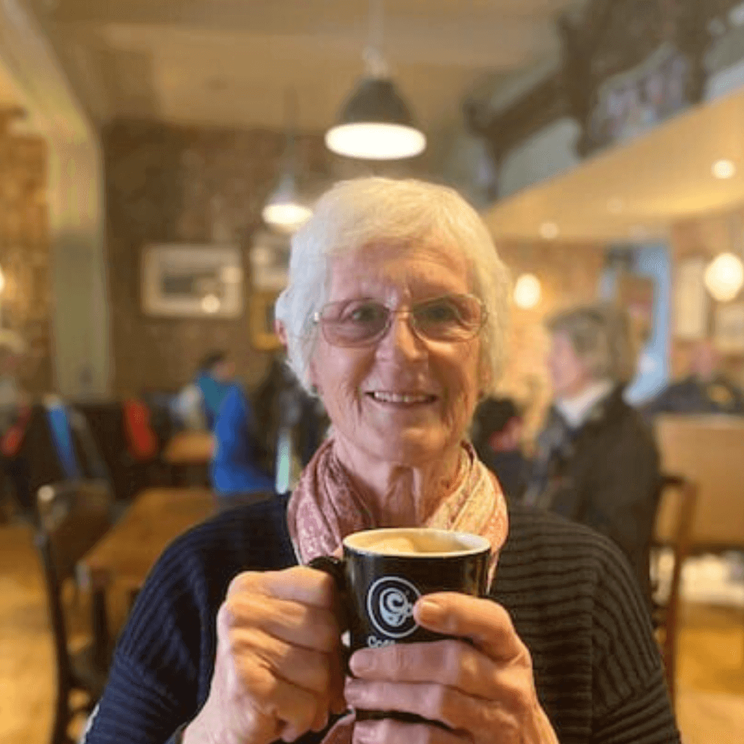 Elderly woman smiling, holding a coffee mug in a cozy cafe setting with soft lighting and blurred background. - Home Instead