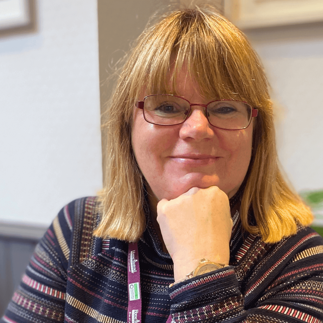 A person with shoulder-length hair and glasses smiles while resting their chin on their hand. - Home Instead