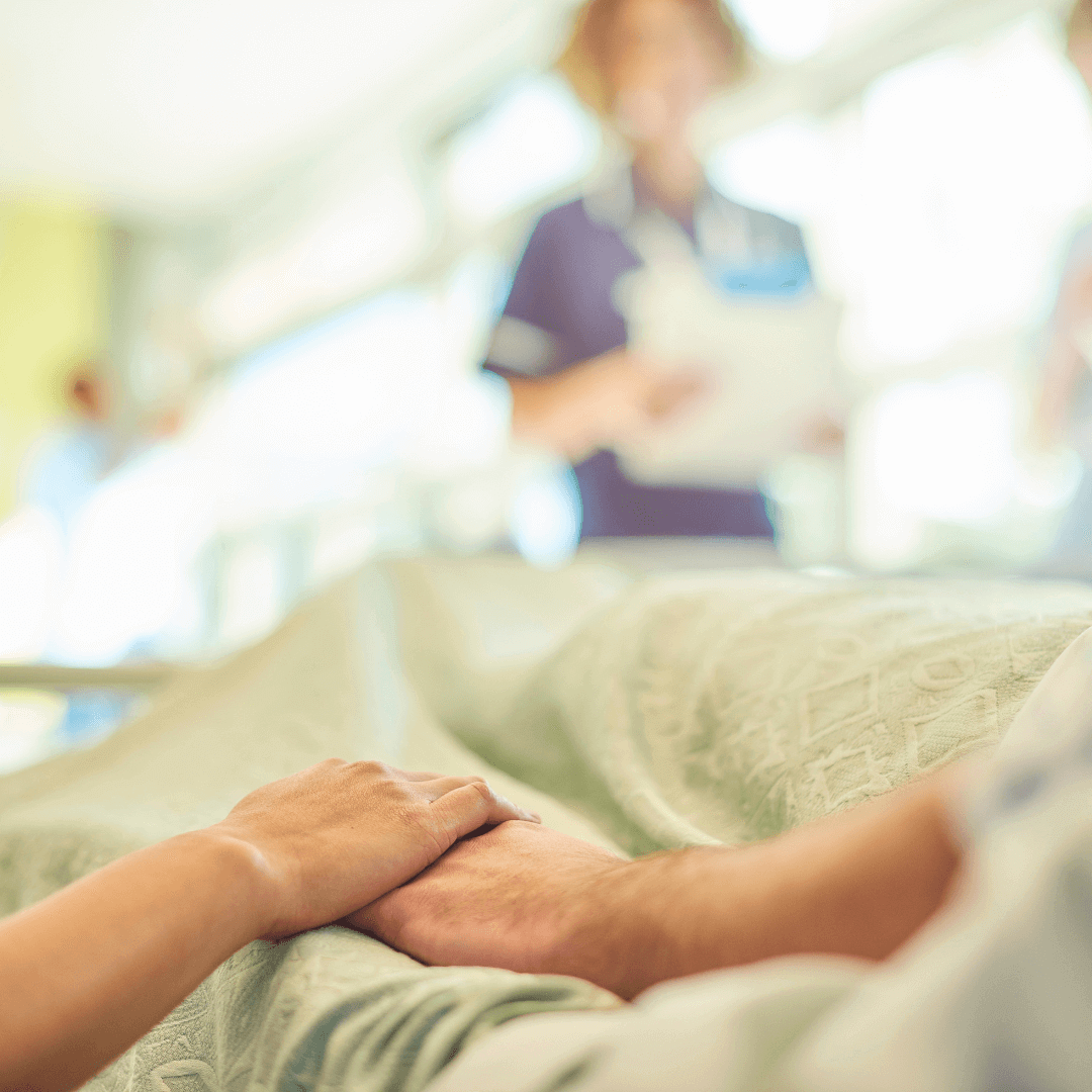 A person holds the hand of someone lying in a hospital bed with a nurse in the background. Caring - Home Instead