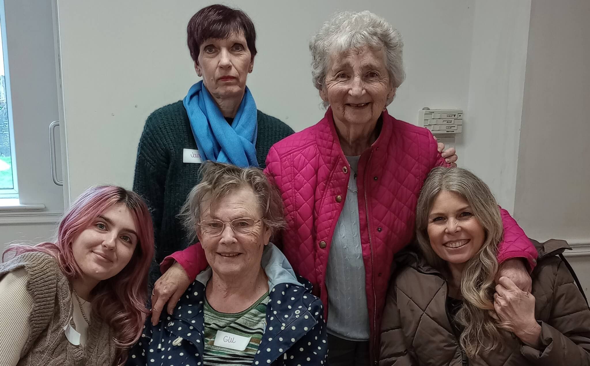 Five women smiling and posing together indoors; two are standing, and three are seated, one in a wheelchair. - Home Instead