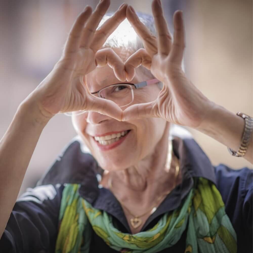 An elderly woman with short gray hair forms a heart shape with her hands, smiling, and wearing glasses. - Home Instead