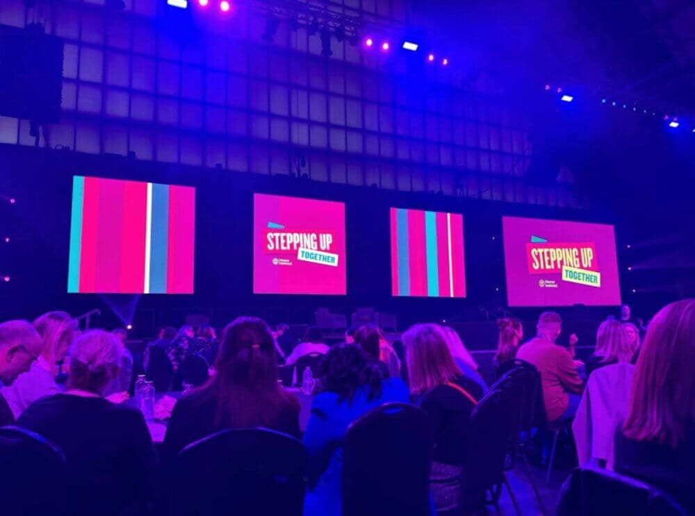 A large audience faces a stage with four colorful screens displaying "Stepping Up Together" in a dimly lit venue. - Home Instead