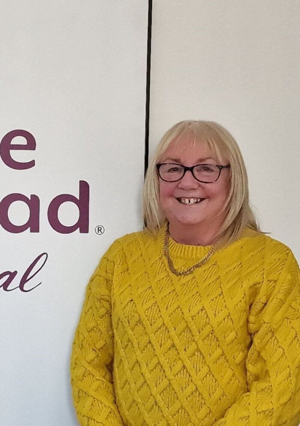 A person wearing glasses and a yellow sweater smiles while standing next to a white backdrop with partial text visible. - Home Instead