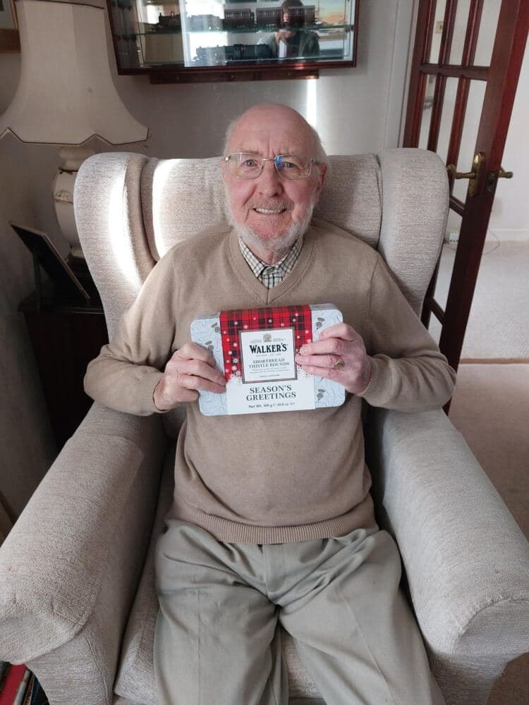 An elderly man sits in an armchair, smiling and holding a box of shortbread cookies. - Home Instead