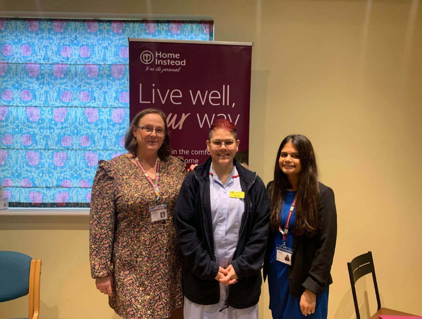 Three women standing in front of a "Home Instead" banner that reads "Live well, your way. - Home Instead