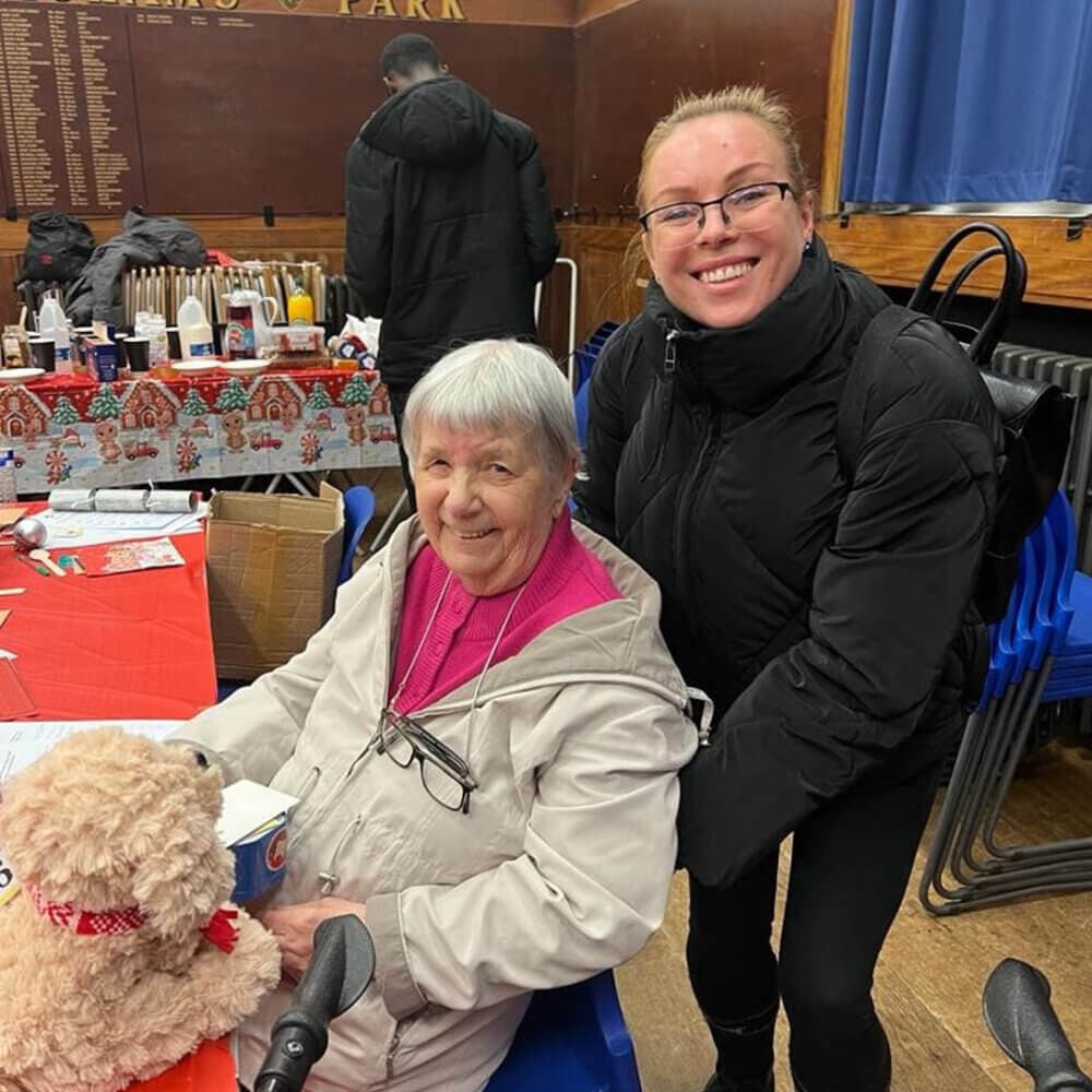 An elderly woman and a younger woman smiling together at a community event, with holiday decorations in the background. - Home Instead