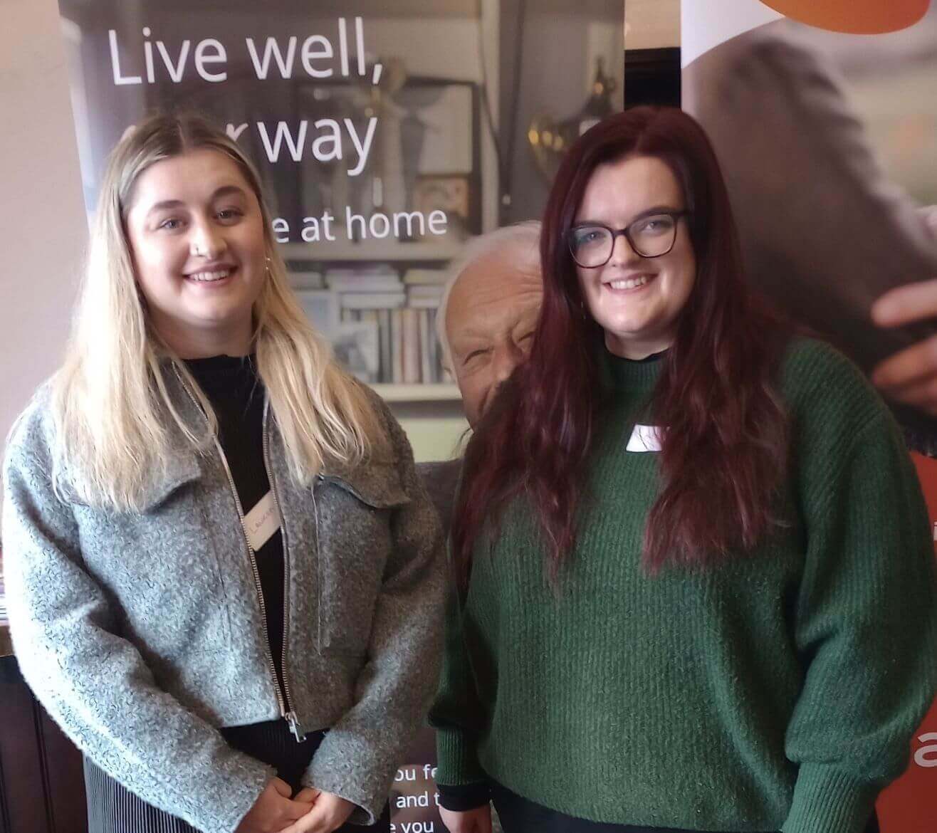 Two smiling women stand side by side in front of a banner that reads "Live well, your way while at home. - Home Instead