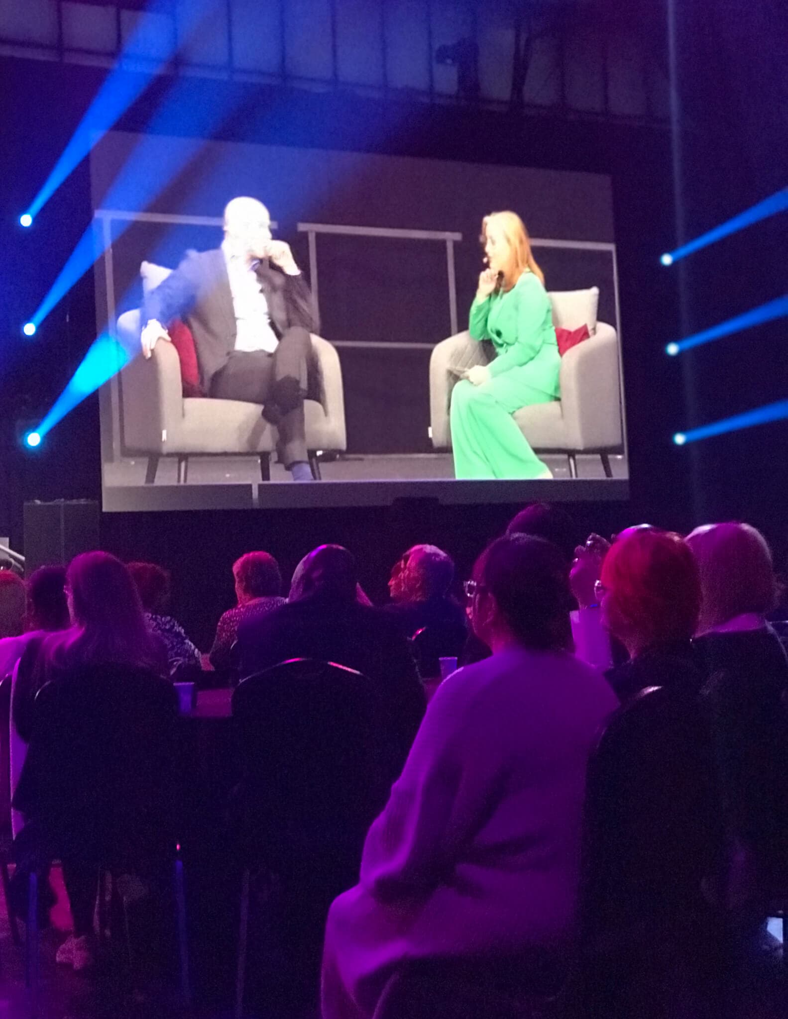 Two people are having a conversation on stage in front of an audience; a large screen displays them. - Home Instead