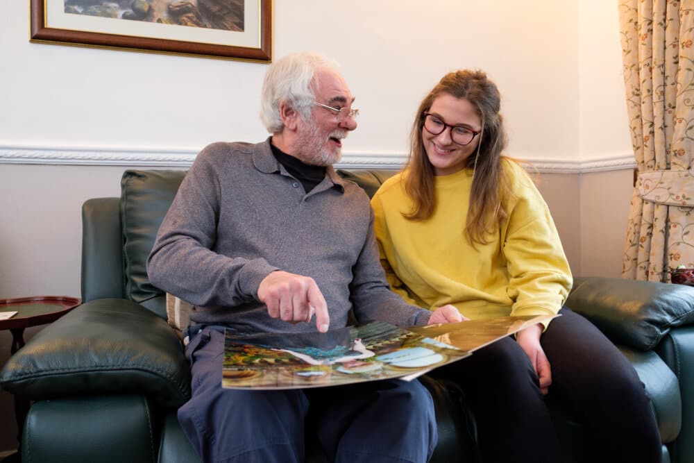 An elderly man and a young woman sit on a couch, smiling and looking at a large photo together. - Home Instead
