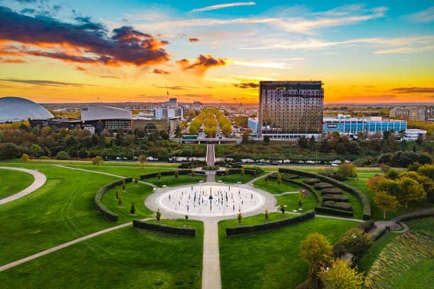 Aerial view of a park with a fountain, surrounded by greenery and buildings, under a vibrant sunset sky. - Home Instead