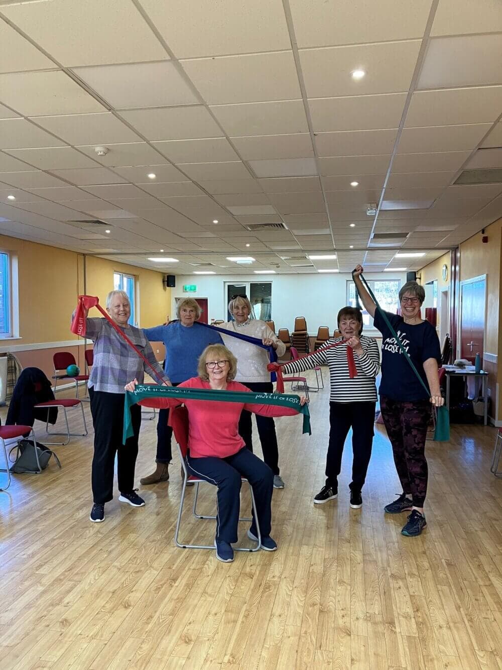 A group of senior women exercising with resistance bands in a bright, spacious room. - Home Instead