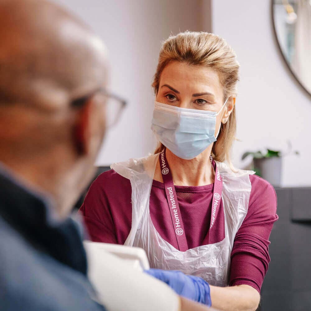 A healthcare worker wearing a mask and apron talks to a seated person, focusing attentively. - Home Instead