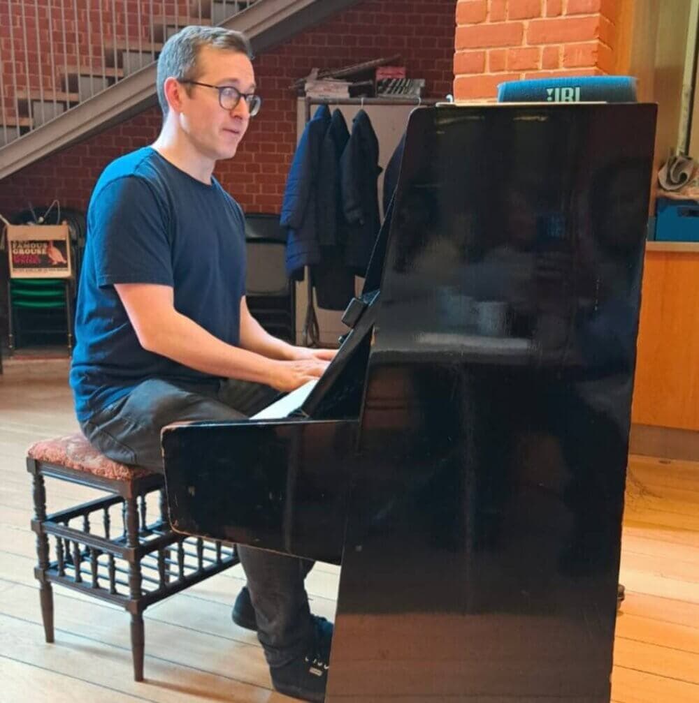 A person wearing glasses plays a piano in a room with brick walls and a staircase in the background. - Home Instead