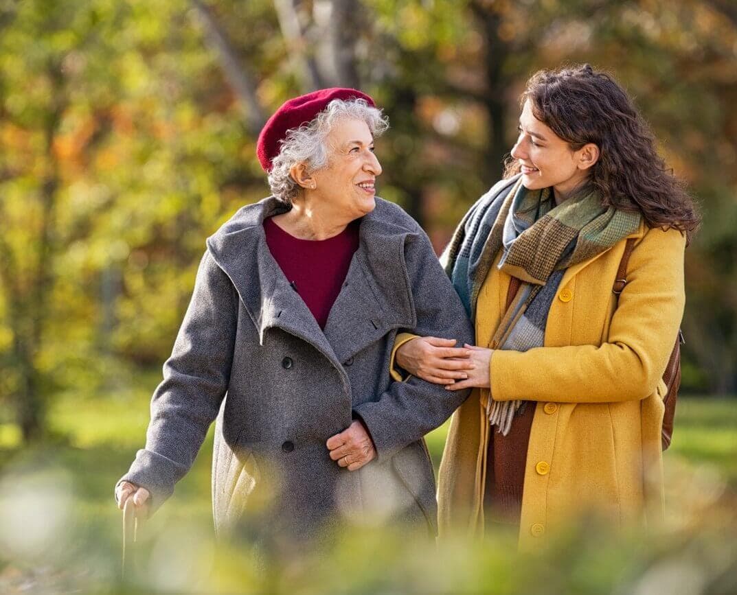 An older woman with a cane and a younger woman, both smiling, walk together in a park on a sunny day. - Home Instead