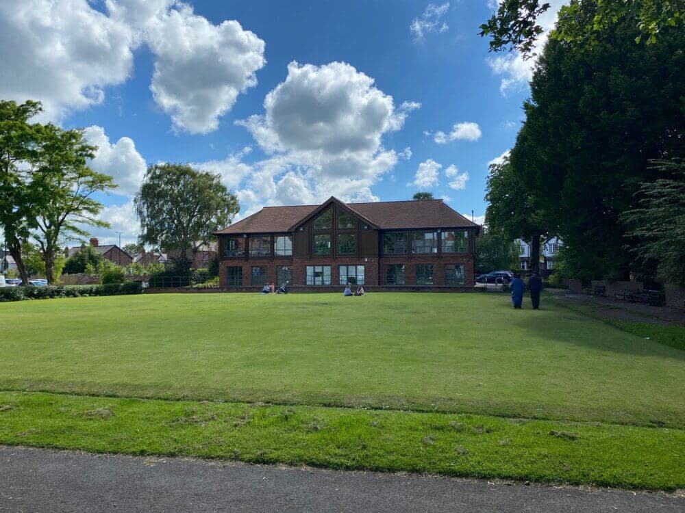 A lush green lawn in front of a large brick building with people sitting and relaxing under a bright blue sky with clouds. - Home Instead