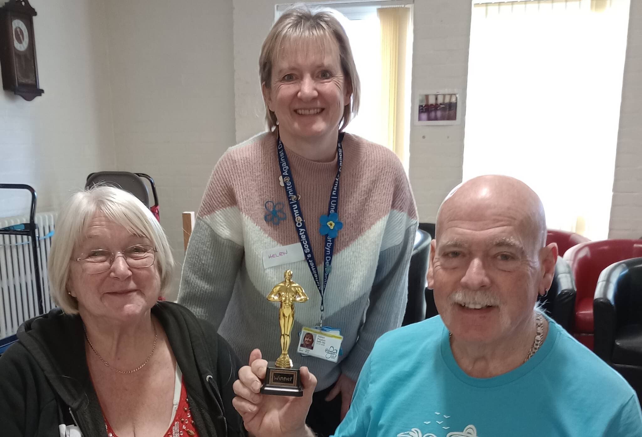 Three smiling people, one holding a small golden trophy; in a cozy room with chairs and a window in the background. - Home Instead