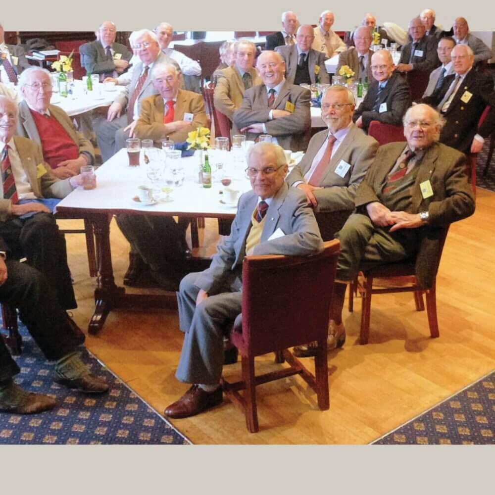 A group of elderly men in suits are gathered around tables in a warmly-lit room, smiling for a group photo. - Home Instead