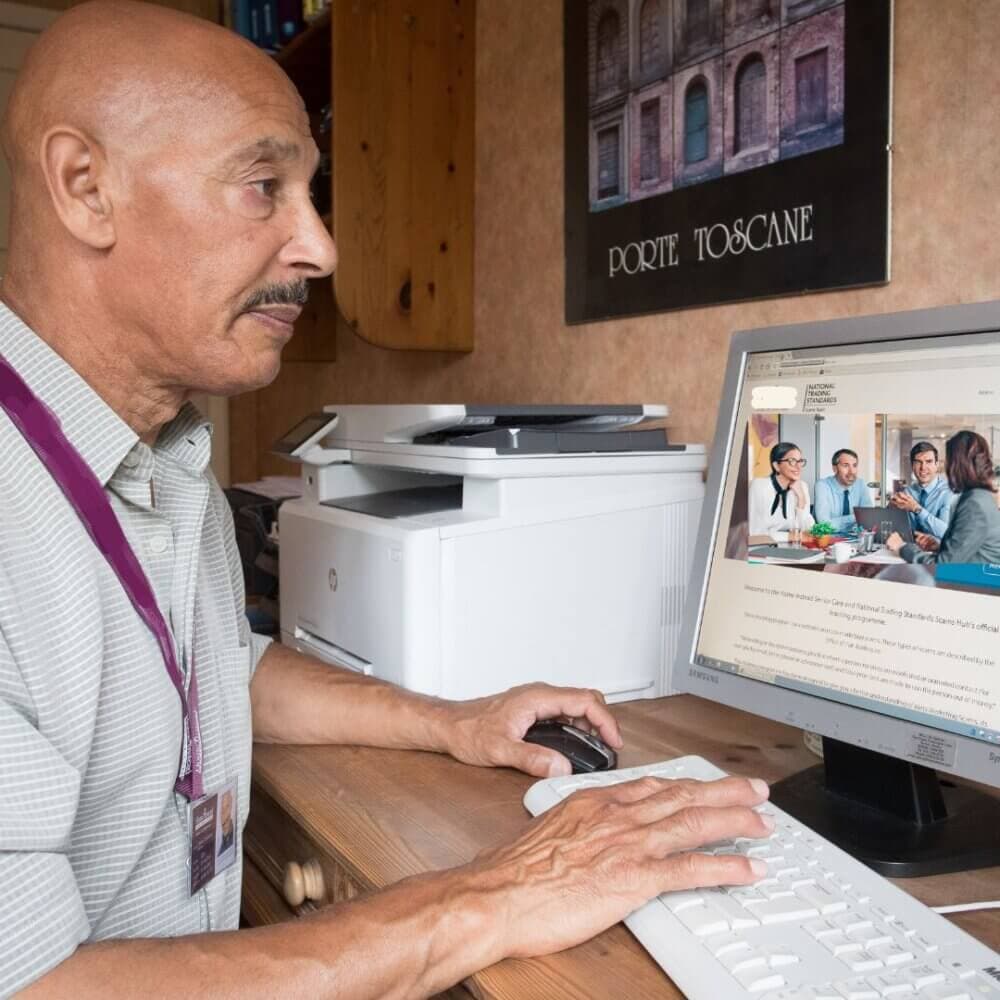 An older man uses a computer at a desk, viewing a webpage with people in a meeting. Printer and wall art in background. - Home Instead