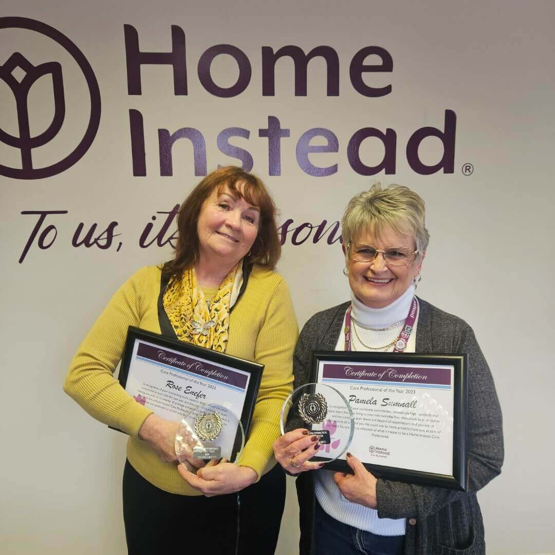 Two women holding certificates and awards in front of a "Home Instead" sign, smiling at the camera. - Home Instead