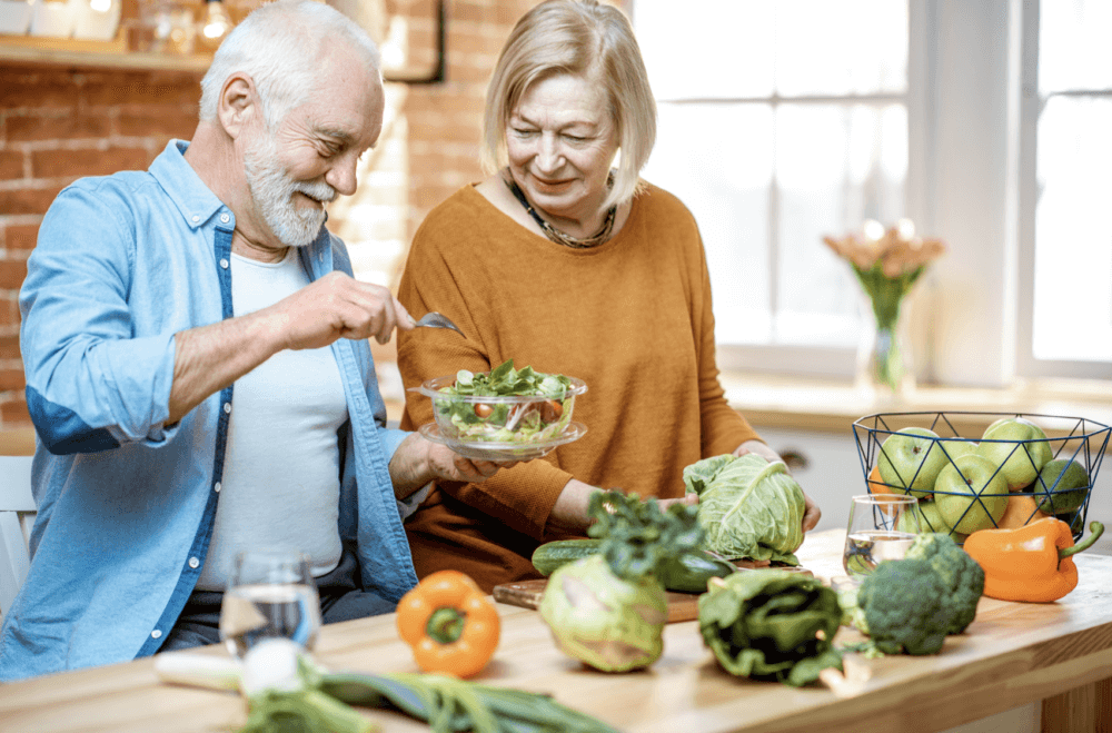 Elderly couple preparing a salad together in a bright kitchen with various fresh vegetables on the table. - Home Instead