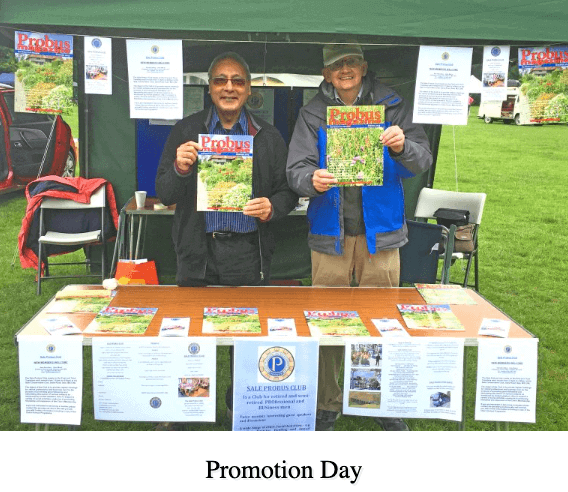 Two people hold magazines at a promotional booth for the Probus Club on a grassy outdoor area. - Home Instead