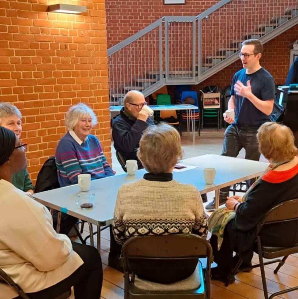 A group of elderly people sit around a table, while a man stands and speaks to them in a community center. - Home Instead