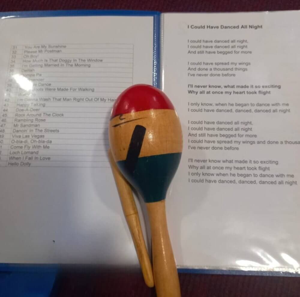 A maraca rests on an open booklet containing song lyrics, with "I Could Have Danced All Night" visible. - Home Instead