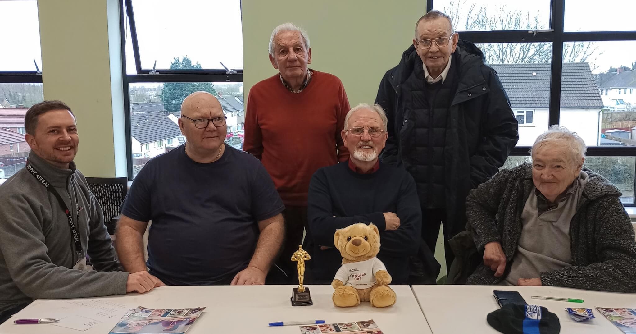 Group of six older adults smiling around a table with a small trophy and stuffed bear in a well-lit room. - Home Instead