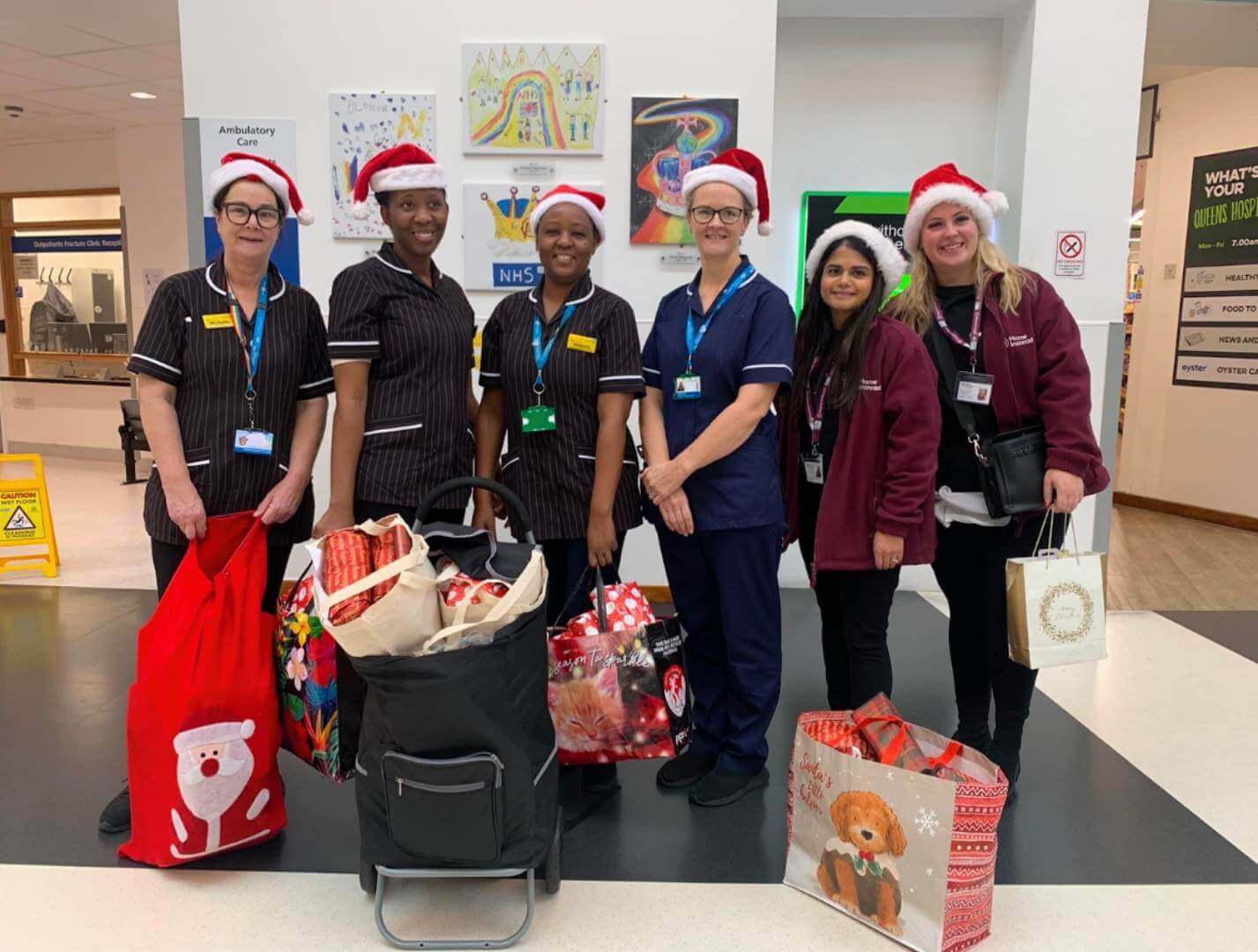 Six hospital staff wearing Christmas hats stand with gift bags in a hospital lobby. - Home Instead