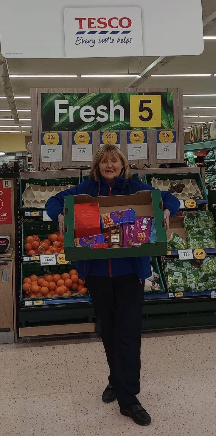 A person holding a box of groceries stands in front of a produce section at Tesco. - Home Instead