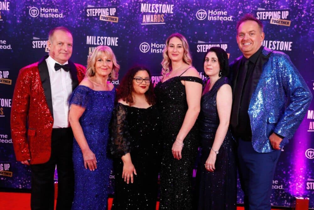 Six people in formal attire posing on a purple starry backdrop at an awards event. - Home Instead