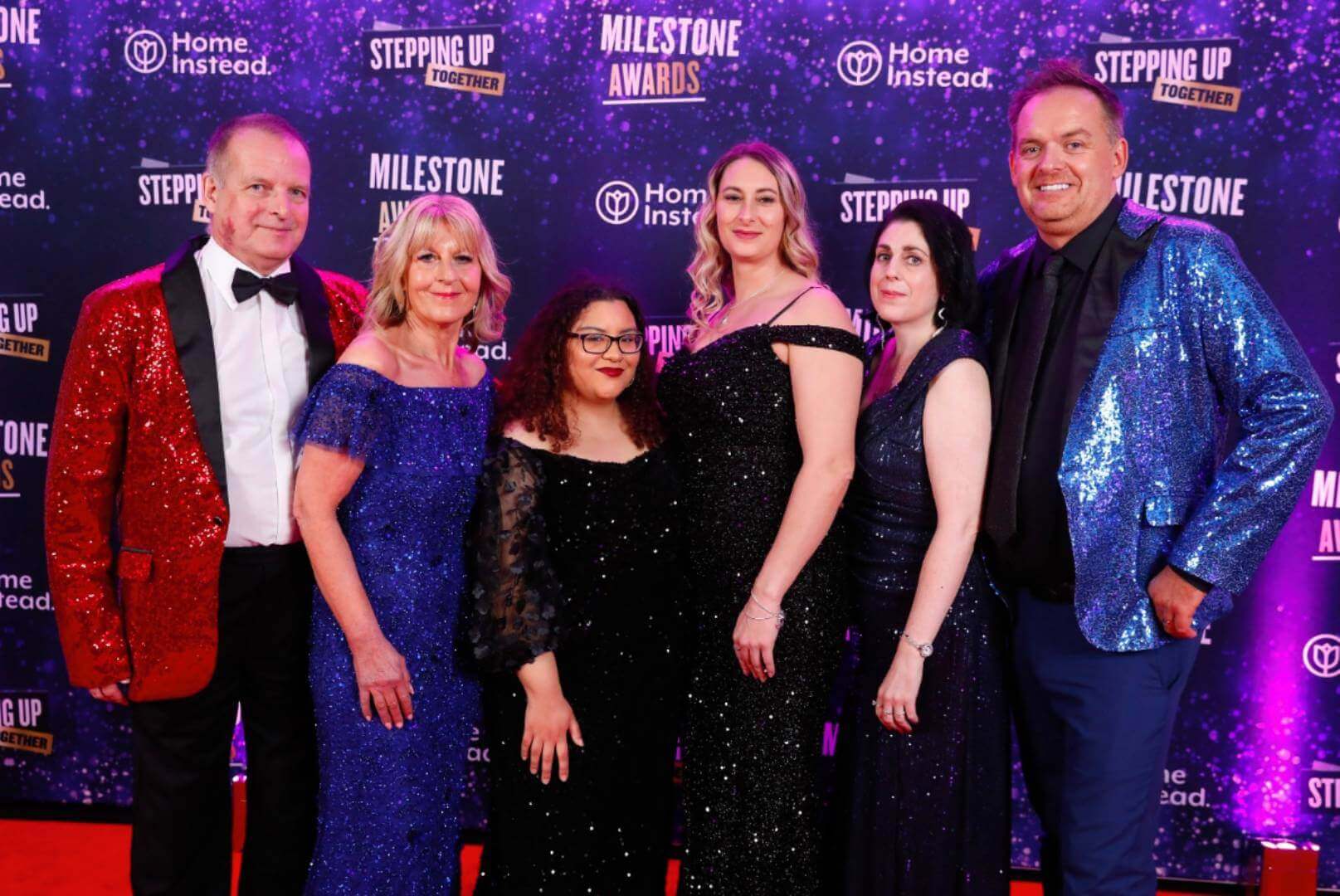 Six people in formal attire posing on a purple starry backdrop at an awards event. - Home Instead