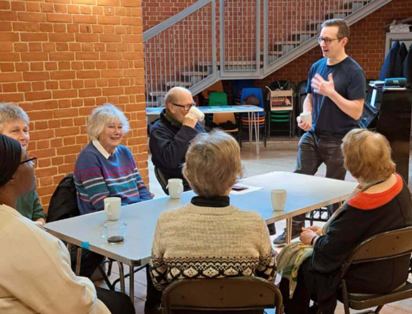 A group of six elderly people sit at a table, listening to a standing man in a casual setting with brick walls. - Home Instead