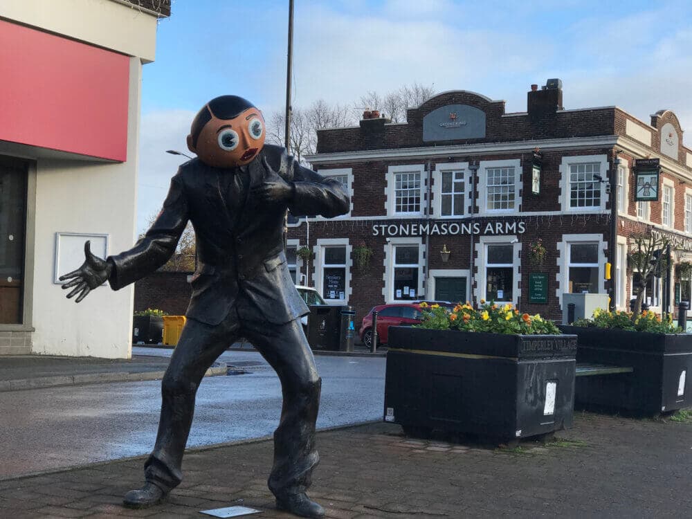 Statue of a person with a large round head in front of a building labeled "STONEMASONS ARMS. - Home Instead