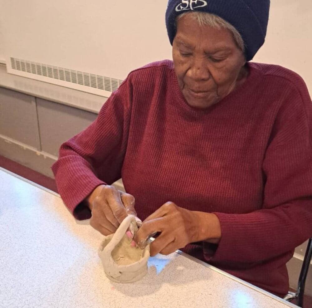 An older woman is making a clay pot at a table, focused on shaping the material with her hands. - Home Instead