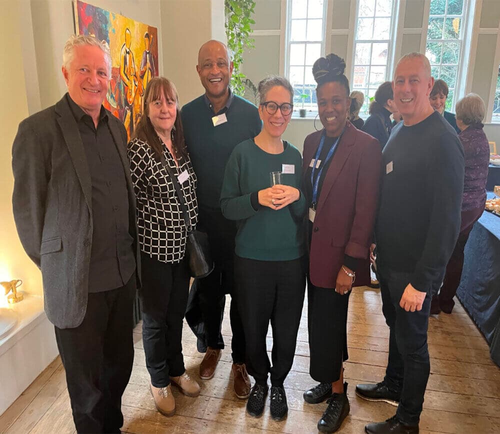 Group of six smiling people standing together in a well-lit room with large windows and artwork on the wall. - Home Instead