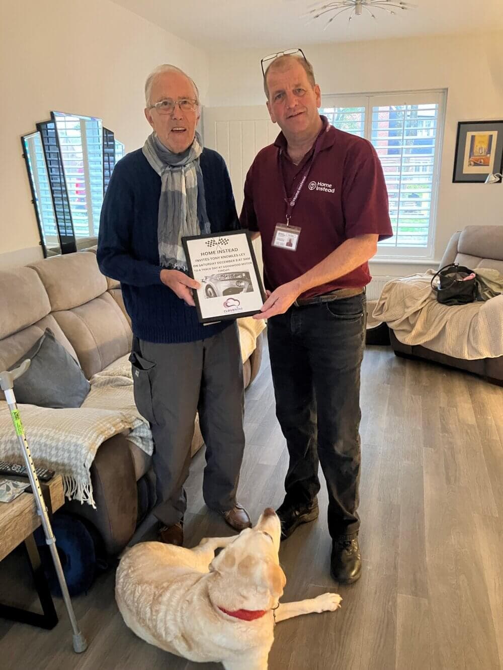 Two men stand in a living room, one holding a certificate, with a dog sitting on the floor in front of them. - Home Instead