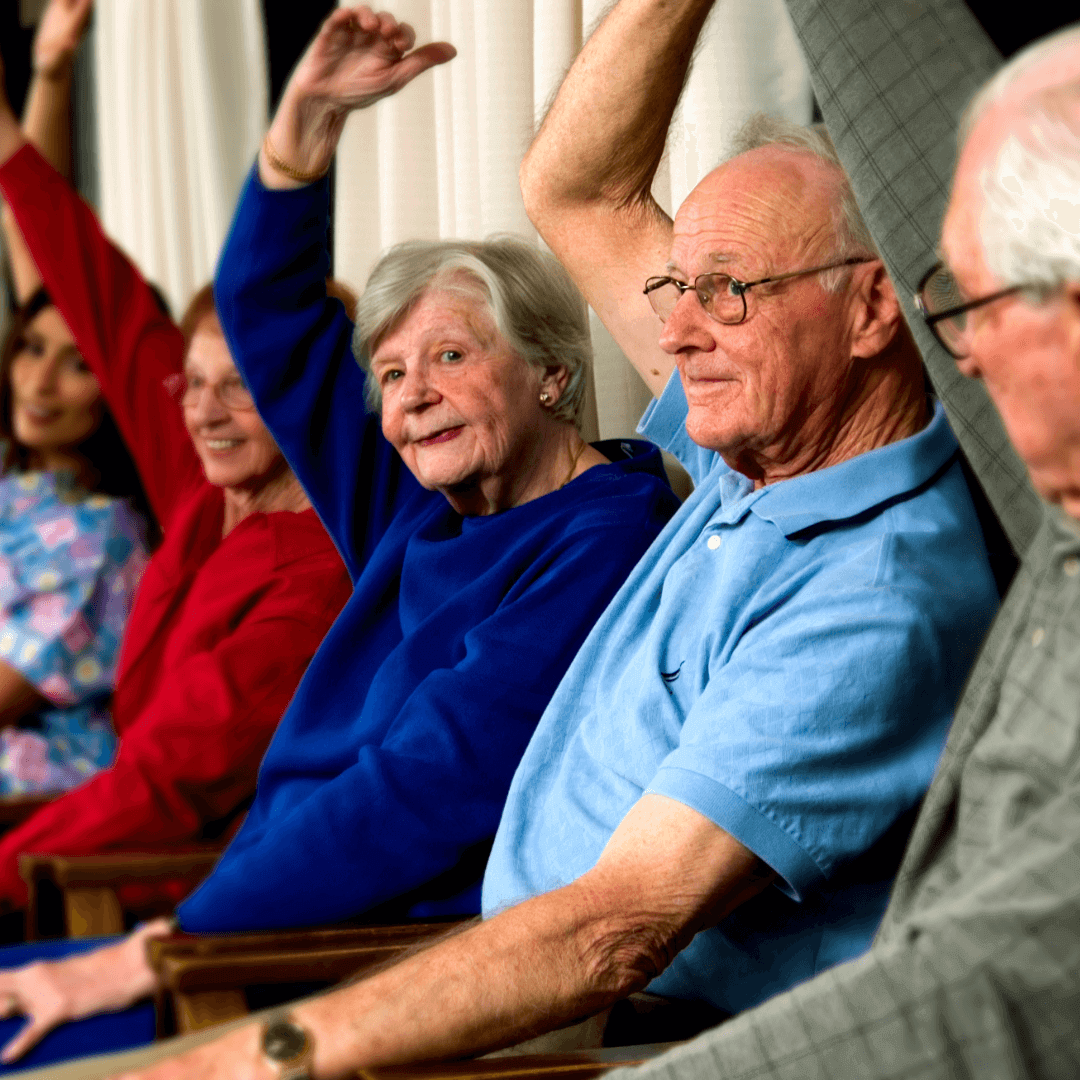 Seniors sitting in chairs, raising their arms during an exercise routine, smiling and engaged. - Home Instead